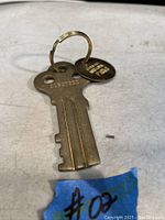 Overall view of the brass Alcatraz key with keyring and attached brass tag on a table with a blue tape labeled "#02".