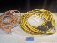 Photo showing two extension cords on a gray surface, one yellow heavy duty 25-foot cord and one orange cord, both with plugs visible.