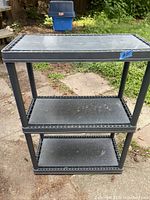 Photo of a heavy plastic shelving unit outdoors showing three black plastic tiers and four legs, exhibiting normal wear.