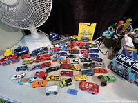 Overview of loose and some boxed Hot Wheels and Mattel cars spread on table next to a fan and scissors holder.