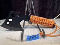 Black sharp axe tool placed on a metal stand, showing sharpened blade with embedded hex wrench cutouts and bottle opener, orange paracord handle with lanyard.