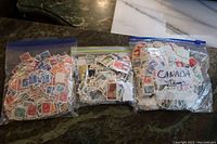 Three clear plastic zip bags each filled with hundreds of used Canadian postage stamps of various colors and designs. One bag labeled 'Canada stamps'.