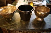 Three flower pots shown side by side on a marble surface: ornate footed brass pot, smooth cylindrical brass pot, and terracotta pot.