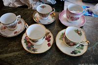 Five different porcelain tea cups and saucers with floral designs and gold trim, including a mustache cup with visible small flake on bottom.
