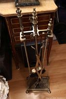 Full view of brass fireplace tool set with tongs, broom, poker, shovel neatly hanging on the stand with a decorative base.