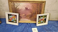 Photo showing two paintings: a larger framed mountain landscape and two smaller floral paintings. Blue tarp background and numbered 46 marker.