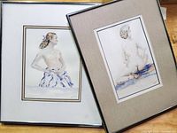 Two framed watercolor paintings of nude women portraits, one seated from behind, one partially nude with cloth, artist signed, displayed tilted against a surface