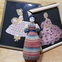 Avon fragrance doll standing in front of two framed silk flower needlework artworks of fashionable ladies, one frame damaged