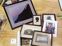 Group of framed photos and prints on table showing varied items including scenic photograph, silhouette art, Kermit photo, historical group photos