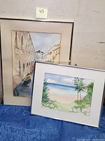 Photo showing two framed watercolor paintings with canal scene and seascape leaning against wall