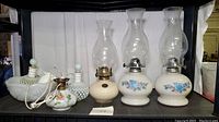 Lot of six vintage glass items: two Fenton hobnail decanters, converted oil lamp, two blue floral hurricane lamps, and white milk glass bowl and vase.