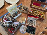Display of costume jewelry including several necklaces, cameo brooch, earrings, watches, and beaded bags in various cases and boxes on a table