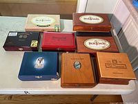 Nine empty cigar boxes of different styles and brands displayed on a table, showing their tops and labels.