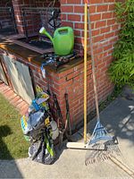 Full view of garden supplies including watering can, rakes, roller, hand tools, and soil bags placed outdoors on a concrete patio near brick wall.