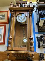 Front view of the wooden case wall clock showing pendulum behind glass door with top ornamental carvings.