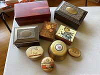 Overview of all eight lidded boxes arranged on a white surface including two large wooden and metal boxes, three small decorative rectangular boxes, and three small oval or round tin boxes with various decorative lids.