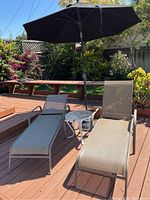 View of patio set with two taupe mesh chaise lounge chairs, small round glass-top table, and large black umbrella on a wooden deck.