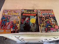 Three vintage comic books in protective sleeves laid on a white surface: Superman's Girl Friend Lois Lane, The Phantom Stranger, and Peter Parker The Spectacular Spider-Man.