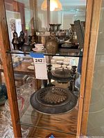 Display cabinet shelf showing assorted metalware items including salt and pepper shakers, brass bell, silverplate baby spoon and food pusher in box, and metal butter dish with perforated lid.