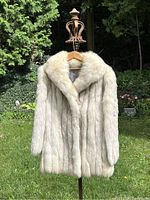 Front view of medium fox fur coat hanging on a wooden hanger in outdoor setting showing full collar and natural cream color.