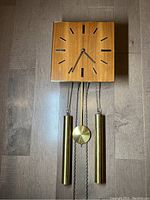 Full view of mid century modern Junghans wall clock with visible teak wood face, black hour markers, brass weights, and brass pendulum on a dark wood floor.