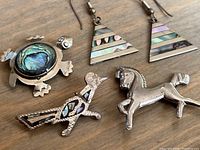 Front view of four silver jewelry pieces: turtle brooch with abalone shell, bird brooch with mother of pearl, horse brooch plain silver, and pair of triangular earrings with mother of pearl inlay.