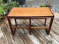 Large teak nesting table with tubular frame and smooth rectangular surface shown on outdoor wood deck.
