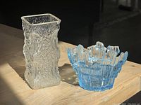 Two vintage glass vases on wooden surface, one clear rectangular textured vase and one blue round vase with jagged rim.