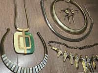 Overview shot of the five jewelry pieces including the green and cream pendant necklace, white segmented collar necklace, gold-tone brooch, textured gold collar, and gold wing design necklace