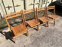 Four wooden slat folding chairs lined up outdoors. Each chair has a slatted seat and back with a foldable wooden frame in natural finish.