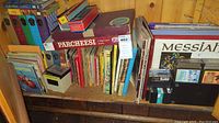 Shelf with various books, board games, cassette tapes, vinyl records, and magazines arranged on wood shelving.