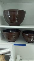 Three brown glazed ceramic bowls on shelf, varying sizes, ribbed design near rim, shiny finish.