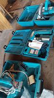 Three teal colored Makita power tools with cords and cases placed on the floor.