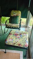 Full view of the metal rocking chair with floral cushion and green pillow, alongside matching ottoman with floral cushion.