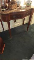 Full view of wooden accent table with one drawer, curved front, tapered legs, and decorative top surface. Table shows signs of wear including scuffs and scratches.
