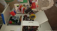 Open cabinet showing multiple cans of paint or related materials, a rubber mallet, packaged items, and cloths on floor nearby