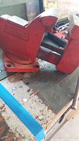 Side angle view showing the overall condition and mounting base of the red metal vise with visible markings.