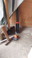 Photo showing metal tamper, orange measuring tape, and portion of broom in corner on concrete floor