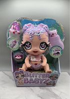 Front view of Mega Glitter Babyz Lila Wildbloom doll in original packaging, showing doll with glittery purple-pink hair, big eyes, floral outfit, and included accessories.