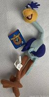 Full view of the multicolor Roadrunner plush toy laying flat on a gray fabric surface, showing front and side details and attached tags.
