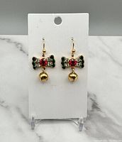 Front view of vintage gold-tone dangle earrings featuring rhinestone bows in green and red, each with a small gold-tone bell charm, displayed on a white earring card against a marble background.