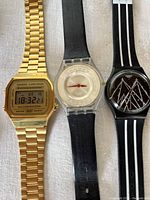 Three watches including Casio gold-tone digital watch, Swatch watch with black band, and Swatch watch with black and white striped band
