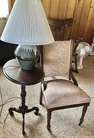 Full view of antique style wooden chair with upholstered seat and back, wooden side table with round top, and ceramic lamp on top with pleated shade.