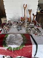 Wide photo showing entire Christmas village with multiple miniature houses and buildings, three wooden reindeers, two silver metallic reindeers, one white reindeer, and Christmas apron laid on table.