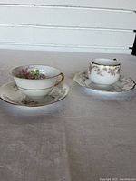 Photo showing two vintage Limose teacups with their matching saucers. One cup has a pronounced floral design and gold handle with a scalloped edge saucer. The other cup also shows pink floral clusters with gold trim on the rim and handle.