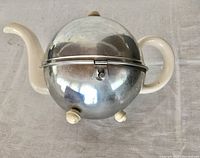 Top view of the teapot showing the cream ceramic spout and handle with metal shell and hinged lid.