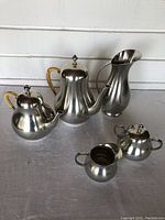 Front view showing all five pewter pieces including teapot, coffee pot, water jug, milk jug, and sugar bowl with distinct wrapped wooden/bamboo handles on teapot and coffee pot, and curved shapes.