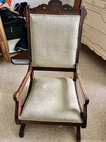 Full front-facing view of the antique rocking chair showing the carved wooden top and cream brocade fabric upholstery on the seat and backrest.