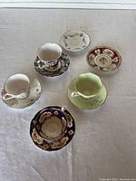 All 5 teacups with their saucers arranged on white background, two small extra plates visible behind