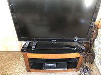 Front view of Samsung TV showing screen and top of stand it rests on, with remote controls and other items placed around it (stand not included).